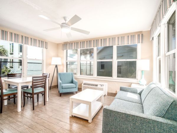 A bright living room with large windows, a ceiling fan, a dining table with chairs, a lounge chair, a sofa, a coffee table, and floor lamps.