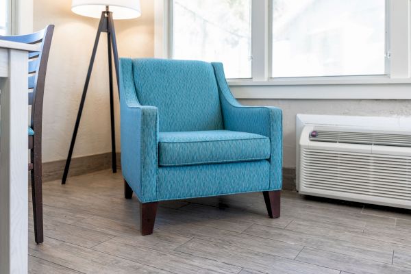 A modern room with a blue armchair, a floor lamp, a table corner, windows providing natural light, and an air conditioning unit on the floor.