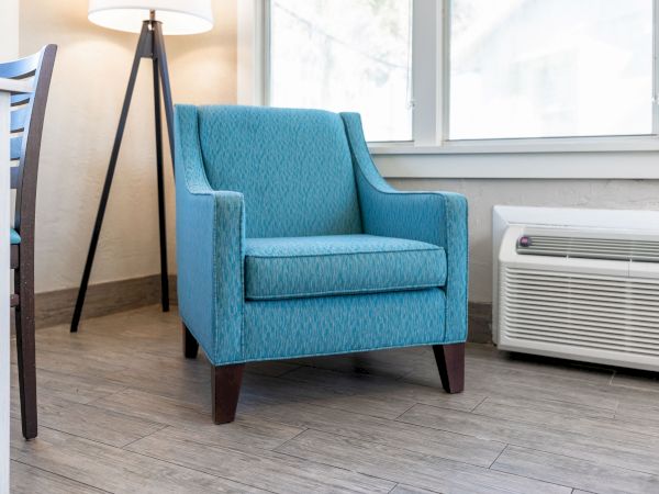 A modern room with a blue armchair, a floor lamp, a table corner, windows providing natural light, and an air conditioning unit on the floor.