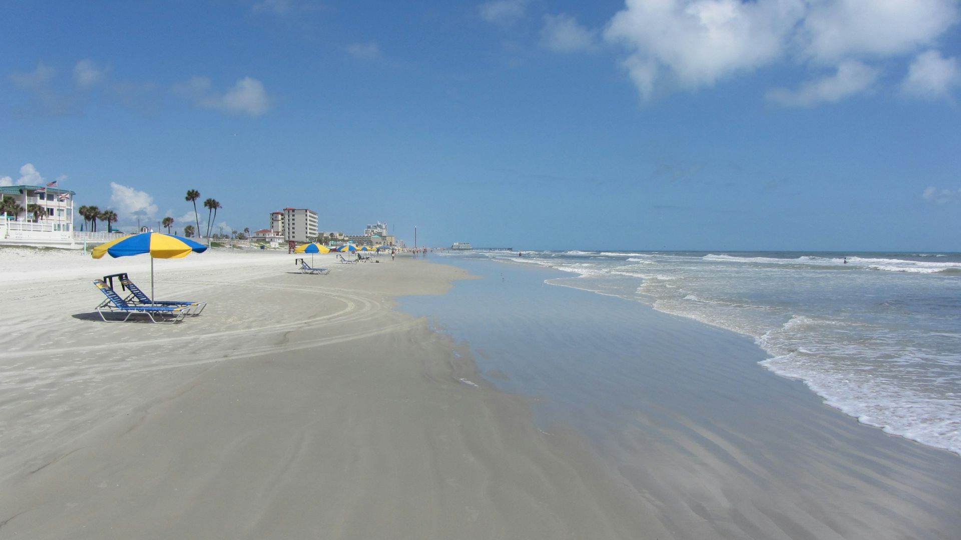 A sandy beach with blue and yellow umbrellas, calm waves, and a clear blue sky with a few clouds.