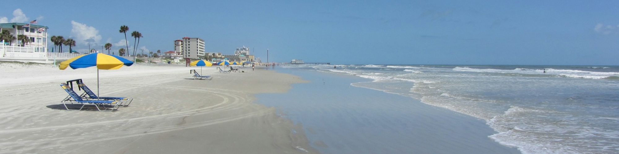 A sandy beach with blue and yellow umbrellas, calm waves, and a clear blue sky with a few clouds.