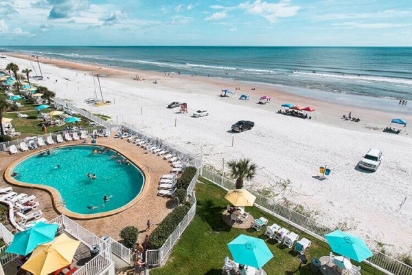 A beach scene with an outdoor pool, lounge chairs, and umbrellas, overlooking the ocean. Vehicles and people are scattered along the sandy shore.