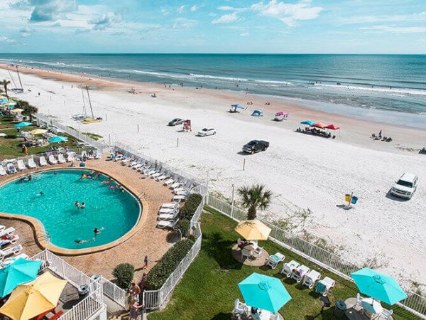 A beach scene with an outdoor pool, lounge chairs, and umbrellas, overlooking the ocean. Vehicles and people are scattered along the sandy shore.
