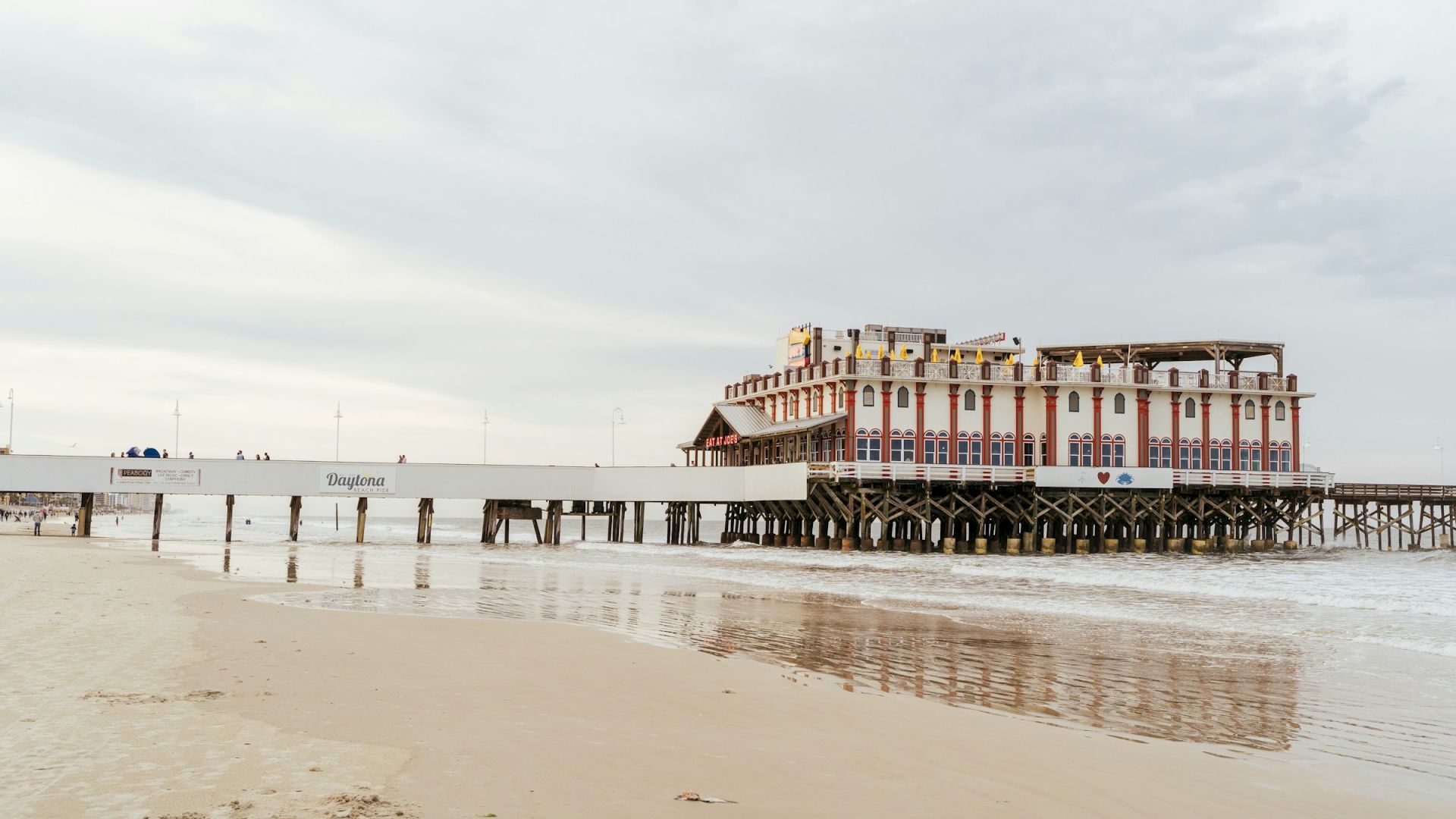 A pier with a large, white and red building on stilts extends over a sandy beach with calm waters under a cloudy sky.