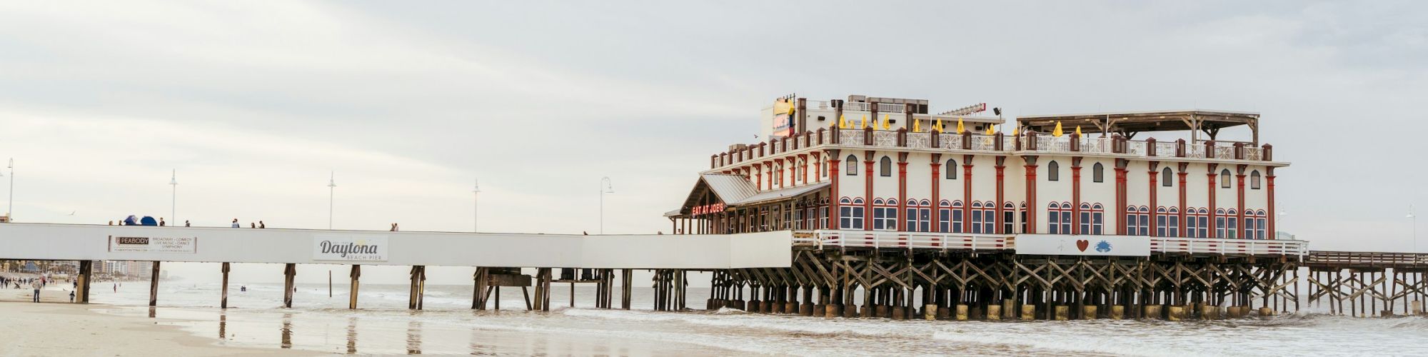 A pier with a large, white and red building on stilts extends over a sandy beach with calm waters under a cloudy sky.