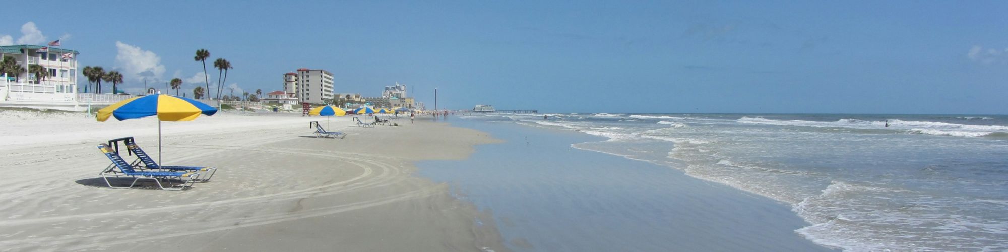 A sandy beach with two beach chairs under yellow and blue umbrellas, waves approaching, and buildings in the background under a blue sky.