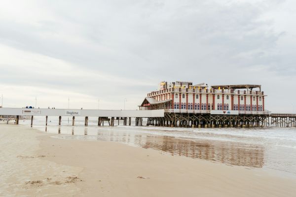 A building on stilts sits over the water, connected to the sandy beach by a long pier on a cloudy day, with reflections visible.