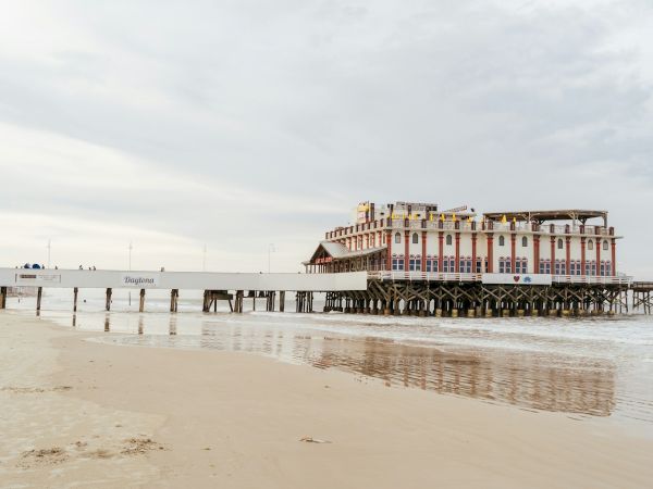 A building on stilts sits over the water, connected to the sandy beach by a long pier on a cloudy day, with reflections visible.