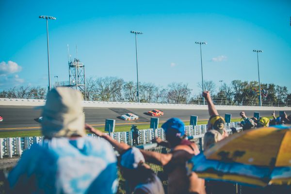 Spectators cheer as cars speed around a racetrack under a clear blue sky, captured in a lively motorsport scene.