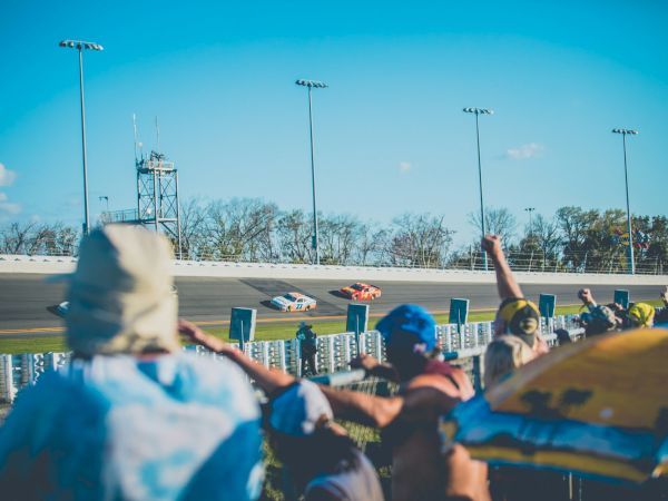 Spectators cheer as cars speed around a racetrack under a clear blue sky, captured in a lively motorsport scene.