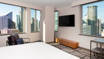 The image shows a modern hotel room with large windows offering a cityscape view, a TV mounted on the wall, and minimalistic furniture.