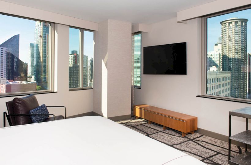 The image shows a modern hotel room with large windows offering a cityscape view, a TV mounted on the wall, and minimalistic furniture.