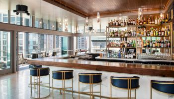 A modern bar with stools, a wide variety of bottled drinks, large windows, and an outdoor seating area with city views, ending the sentence.