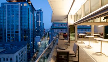 A modern urban balcony at dusk, featuring seating and tables, overlooks a city street and tall buildings.