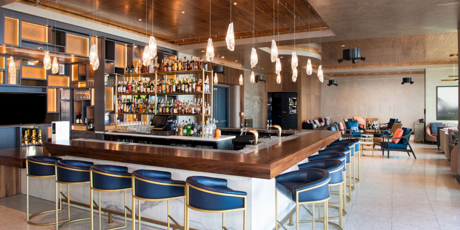 A modern bar with stylish blue stools, hanging pendant lights, a well-stocked shelf of bottles, and a spacious, cozy seating area.