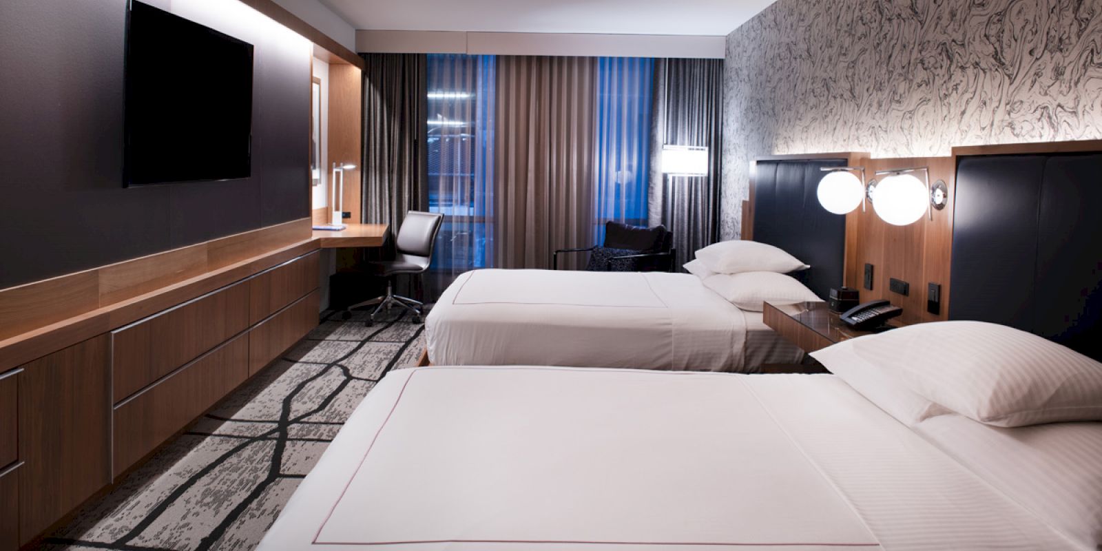 The image shows a modern hotel room with two neatly made beds, a large flat-screen TV, a desk with a chair, and stylish wall decor.