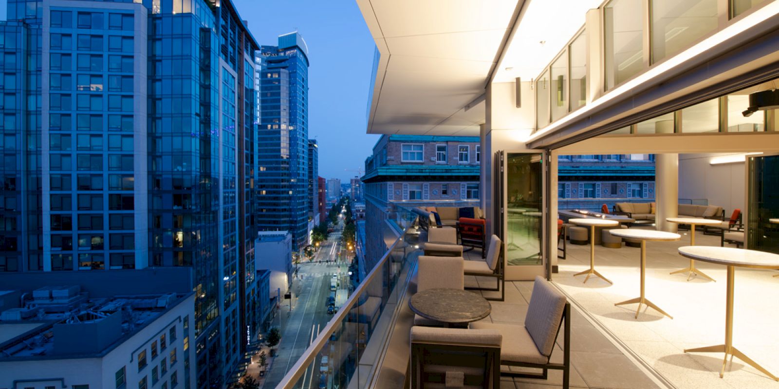 A modern balcony with seating and tables overlooks a city street filled with tall buildings under the evening sky, all lit by streetlights.