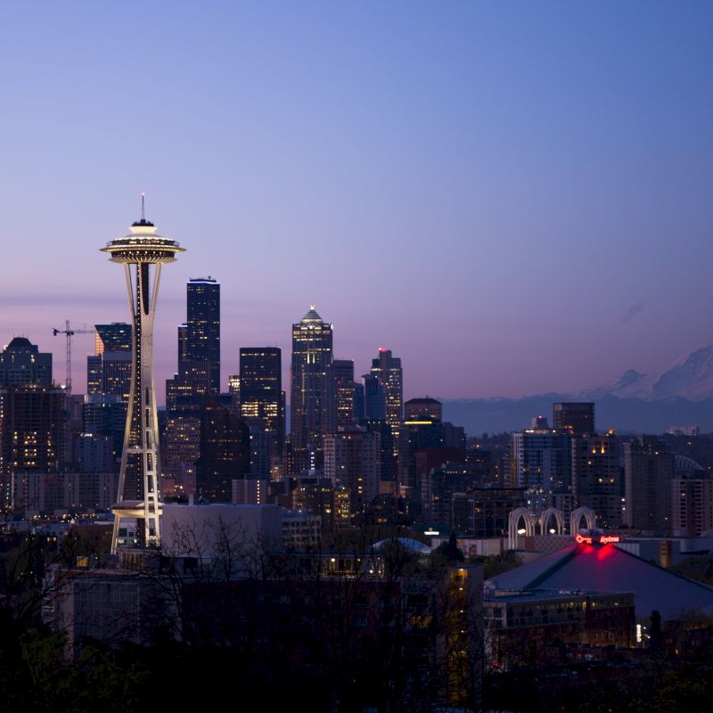 A cityscape of Seattle at dusk, featuring the iconic Space Needle, surrounding buildings, and Mount Rainier visible in the background.