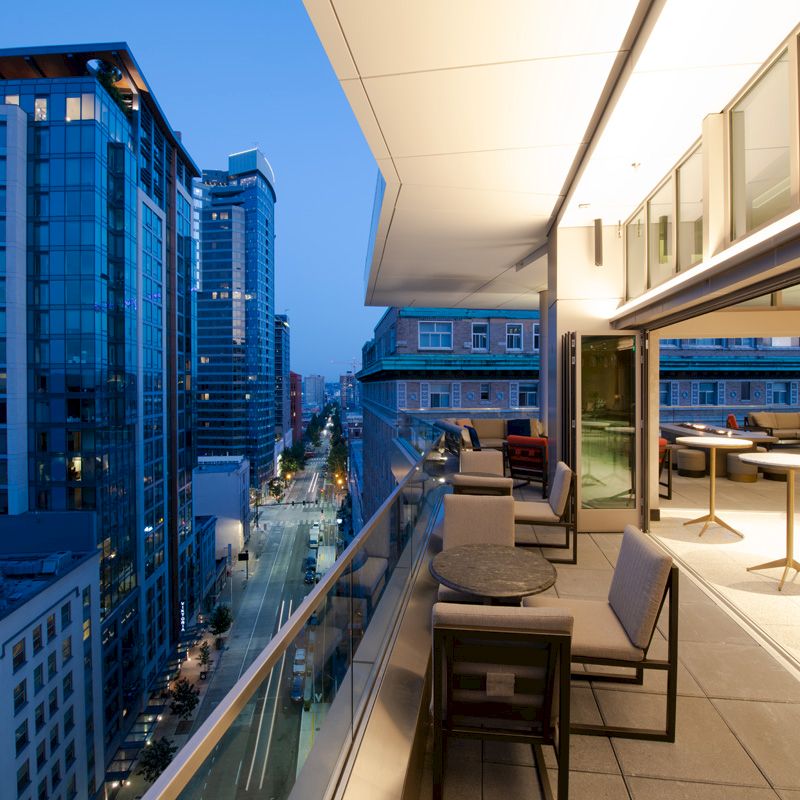 A modern cityscape with high-rise buildings, viewed from a balcony with tables and chairs, during twilight.