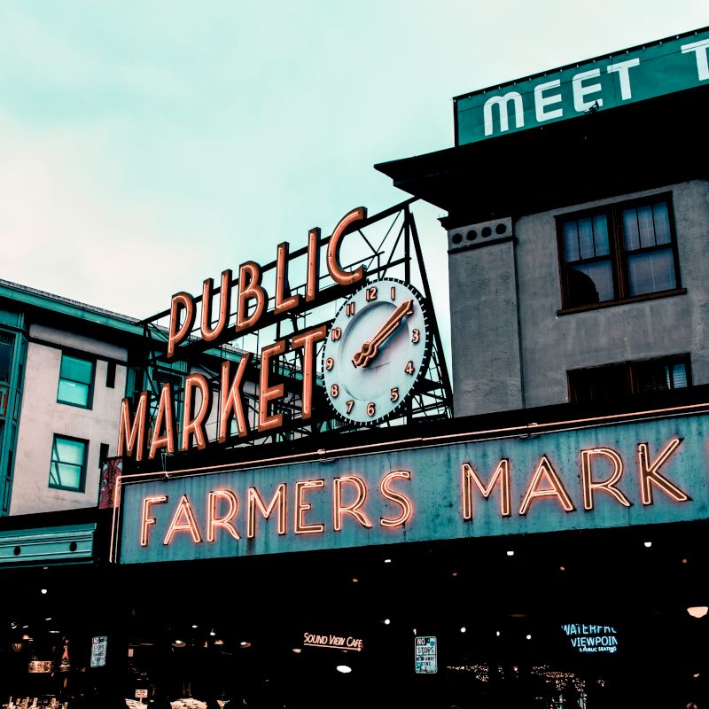 The image shows the entrance to a public market with a neon sign reading "FARMERS MARKET" and a clock. There's also a building with a sign saying "MEET THE."