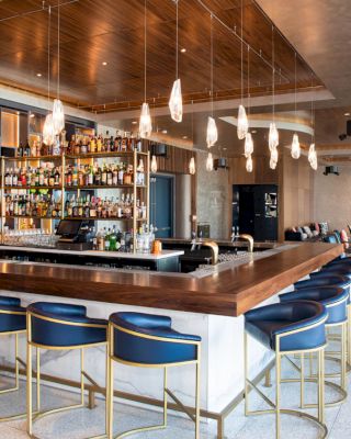 The image features a modern bar with blue chairs, a wooden countertop, and a variety of bottles on shelves behind. Suspended lights hang above.