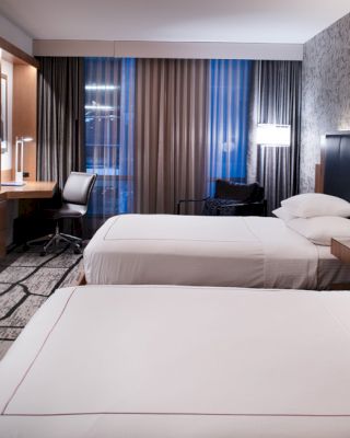 The image shows a modern hotel room with two neatly made beds, a flat-screen TV, a work desk, and patterned wallpaper on the accent wall.