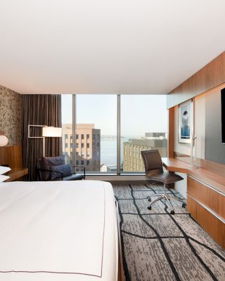 A modern hotel room with a large bed, wall-mounted TV, desk, chair, and a view of city buildings through a wide window.