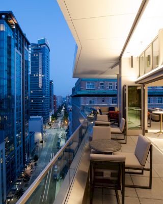 A modern balcony view with seating areas overlooks a cityscape with tall buildings and streets, revealing an urban setting as evening falls.
