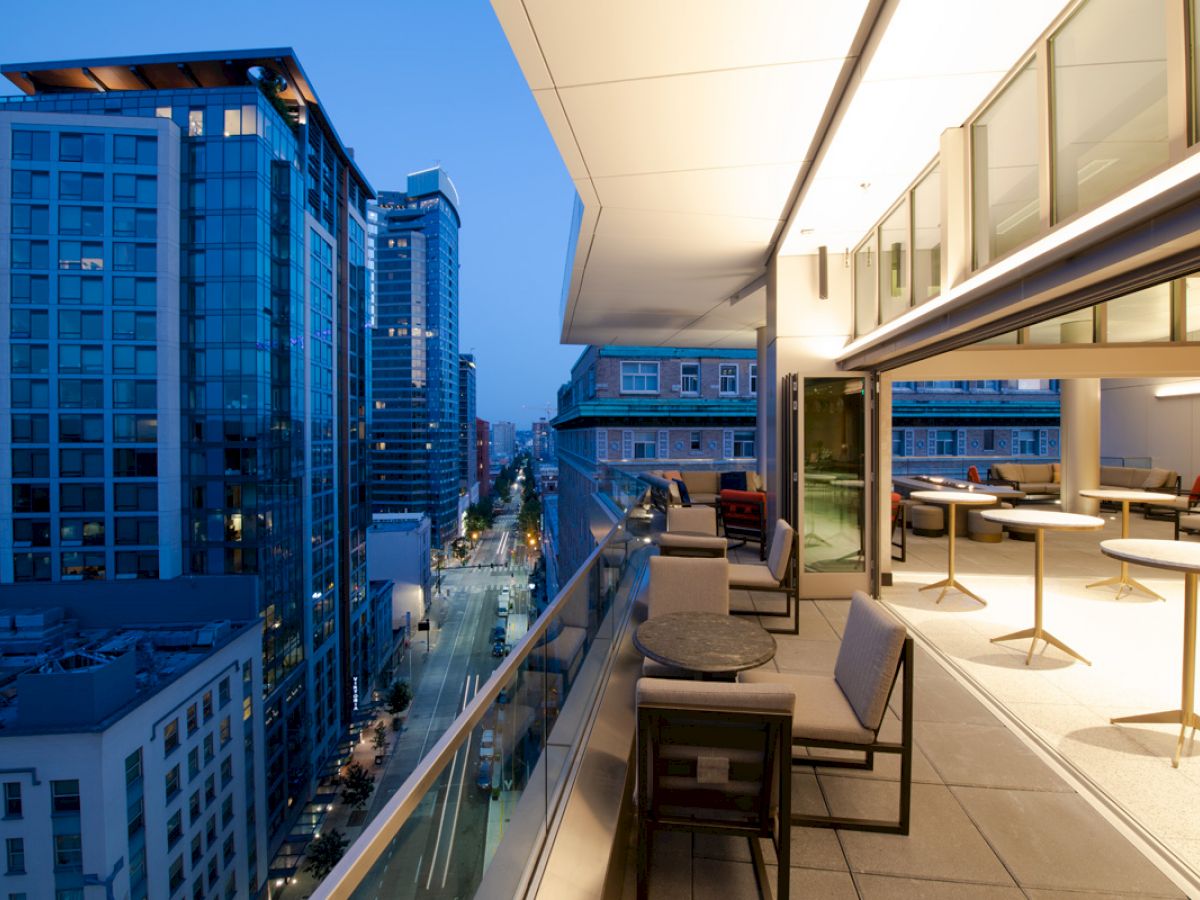 A modern urban balcony with seating and tables overlooks a cityscape with tall buildings and a lit street at dusk.