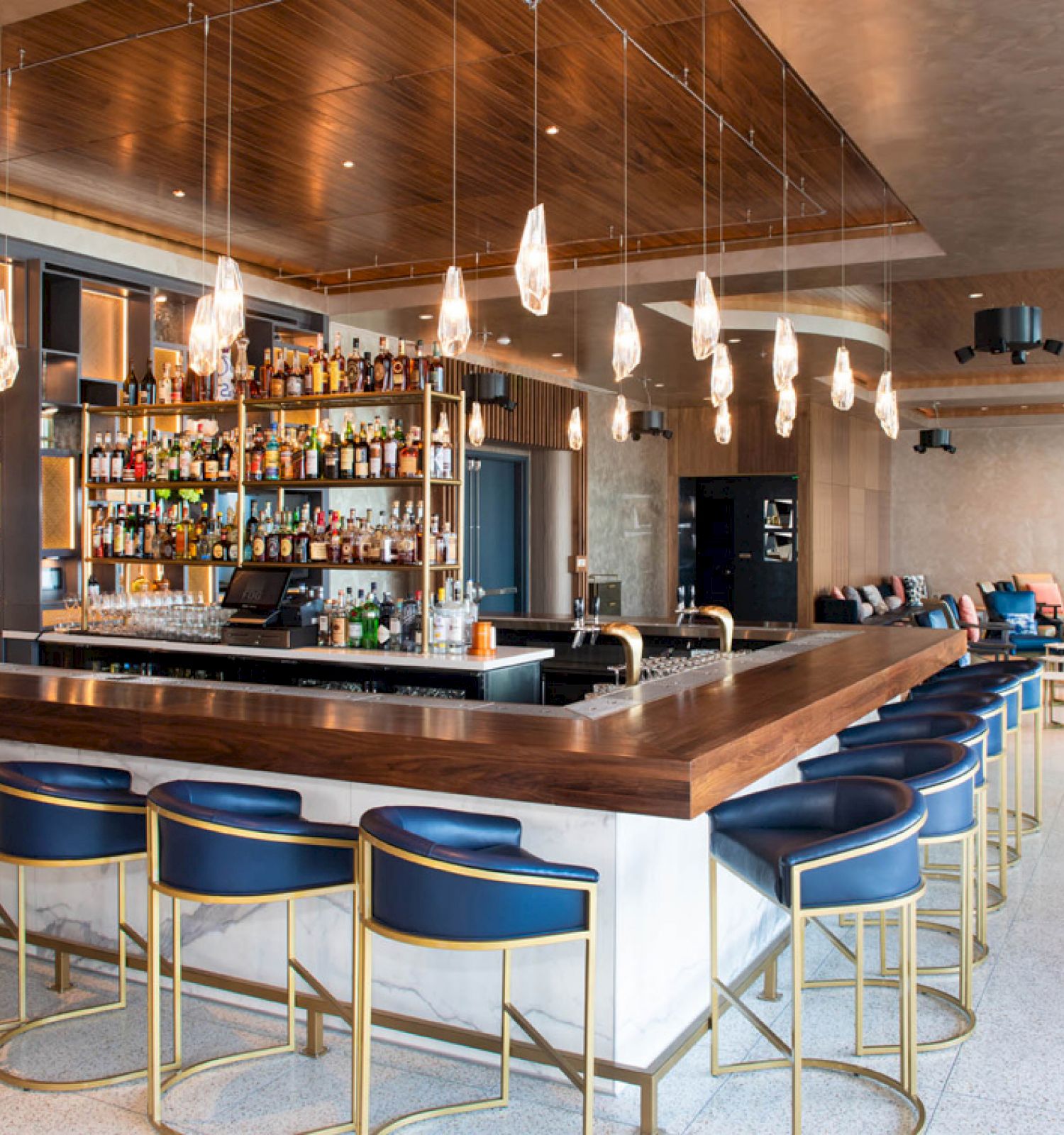 A modern bar with a wooden counter, blue stools, hanging lights, and a well-stocked shelf behind it in a stylish lounge setting.