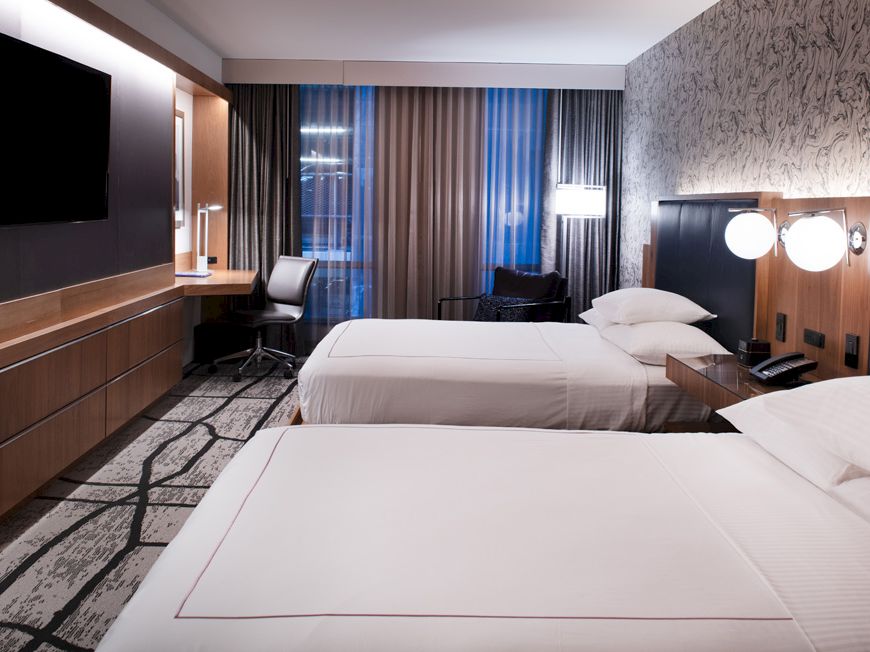 A modern hotel room features two double beds, a wall-mounted TV, a work desk with a chair, and large windows with curtains, all in neutral tones.