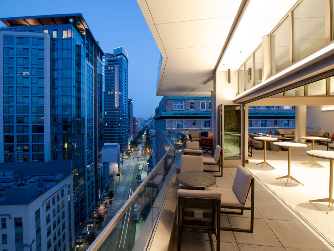 A modern cityscape at dusk with a view from a balcony featuring outdoor seating, high-rise buildings, and a partially open indoor area.
