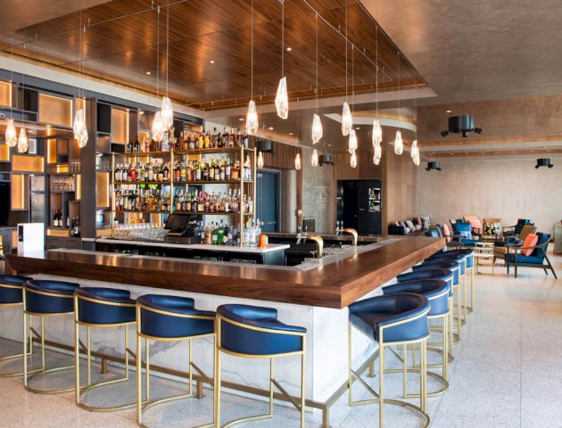 A modern bar with a wooden counter, blue and gold stools, and hanging pendant lights, featuring a well-stocked back bar and seating areas.