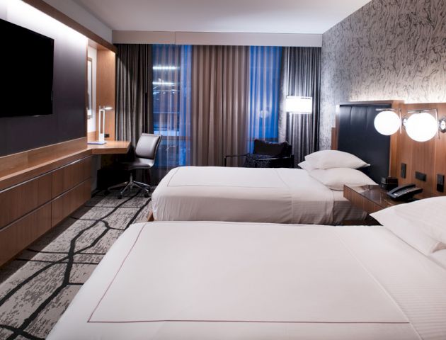 A modern hotel room with two neatly made beds, a large TV, desk with a chair, and stylish decor including patterned wallpaper and curtains ending the sentence.