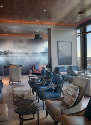 The image shows a modern lounge with comfortable seating, large windows offering a scenic view of a waterfront, and a mural depicting a skyline.