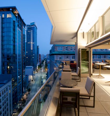A modern balcony with tables and chairs overlooks a city street lined with tall buildings in the evening.