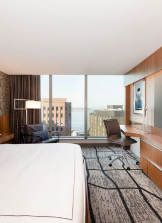 The image shows a modern hotel room with a king-sized bed, a large TV, a desk and chair, and large windows offering a city view.