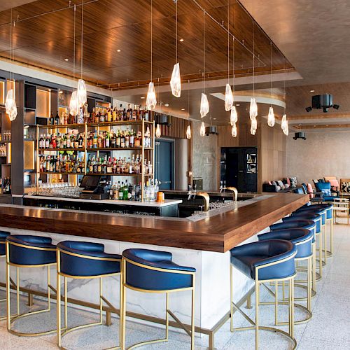 The image shows a modern bar with a wooden counter, blue cushioned seats, and hanging pendant lights. Bottles and glasses are arranged on shelves.