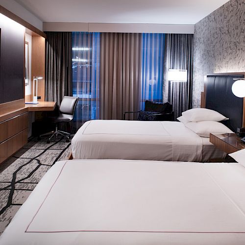 A modern hotel room with two beds, a TV, a desk, and large windows with curtains, featuring contemporary decor and a clean, minimalist design.
