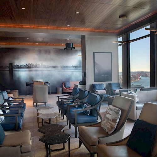 The image shows a modern lounge area with multiple comfortable chairs, large windows offering a scenic view, and a mural on the back wall.