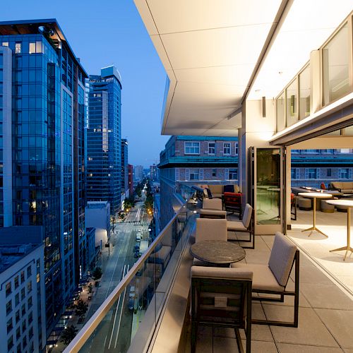 A modern balcony with chairs and tables overlooks a cityscape of tall buildings during twilight, featuring illuminated streets and glass structures.