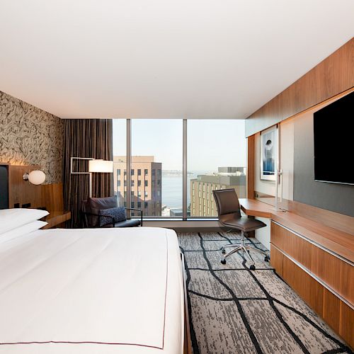 The image shows a modern hotel room with a large bed, a desk with a chair, a flat-screen TV, and a view of buildings and water through large windows.