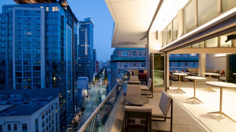 The image shows a modern cityscape view from a high-rise balcony with outdoor seating and tables. The city buildings are illuminated at dusk.