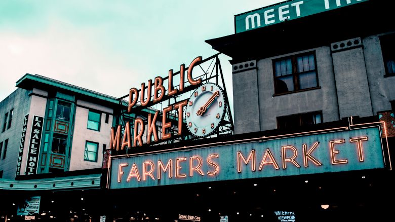 The image shows the iconic neon sign of a public market, advertising a farmers market, with a clock and surrounding buildings in the background.
