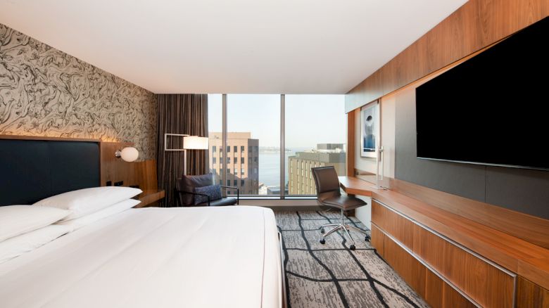 A modern hotel room features a large bed, a work desk with a chair, a flat-screen TV, and floor-to-ceiling windows with a city view, ending the sentence.