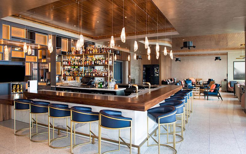 A modern bar with a sleek counter, blue and gold stools, hanging lights, and a well-stocked liquor display; seating area visible in the background.