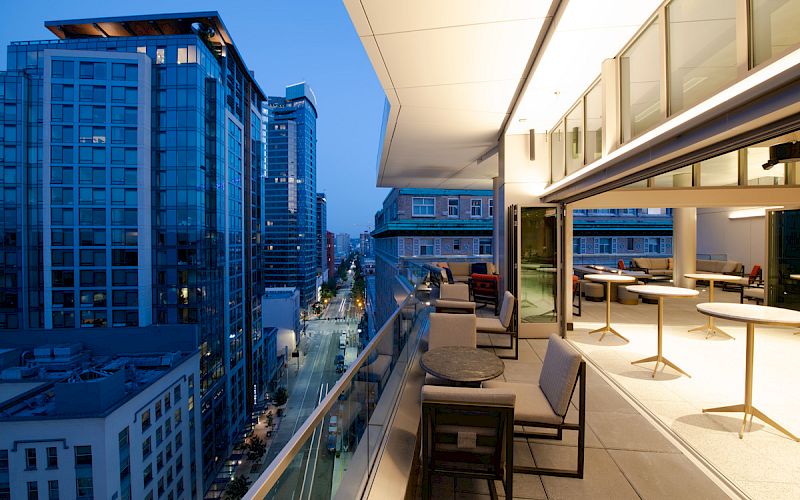 A modern balcony with seating overlooks a city street lined with tall buildings during dusk, with indoor and outdoor seating areas visible.