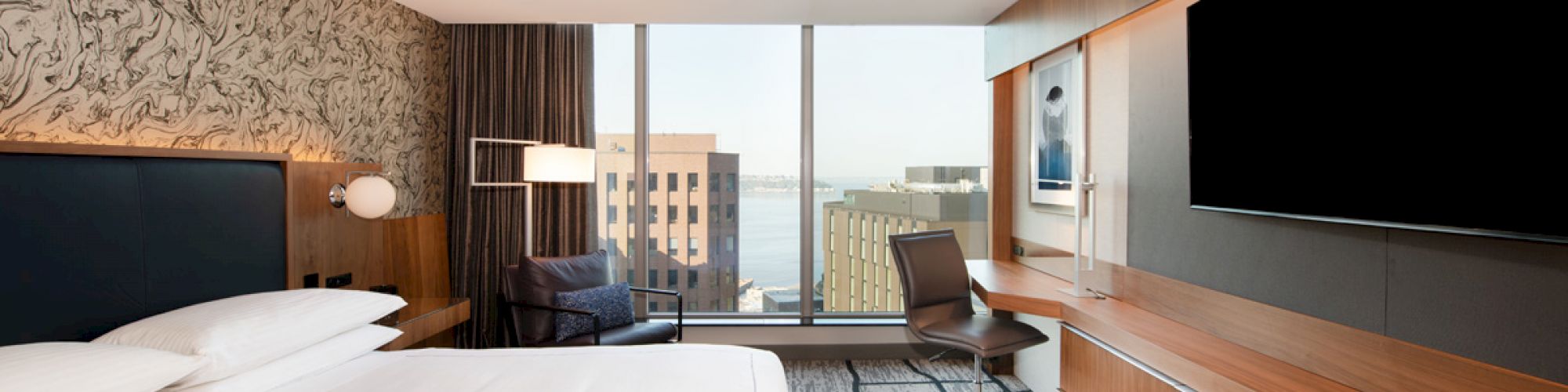A modern hotel room with a large bed, wall-mounted TV, desk, chair, armchair by the window, and a view of city buildings and water outside.