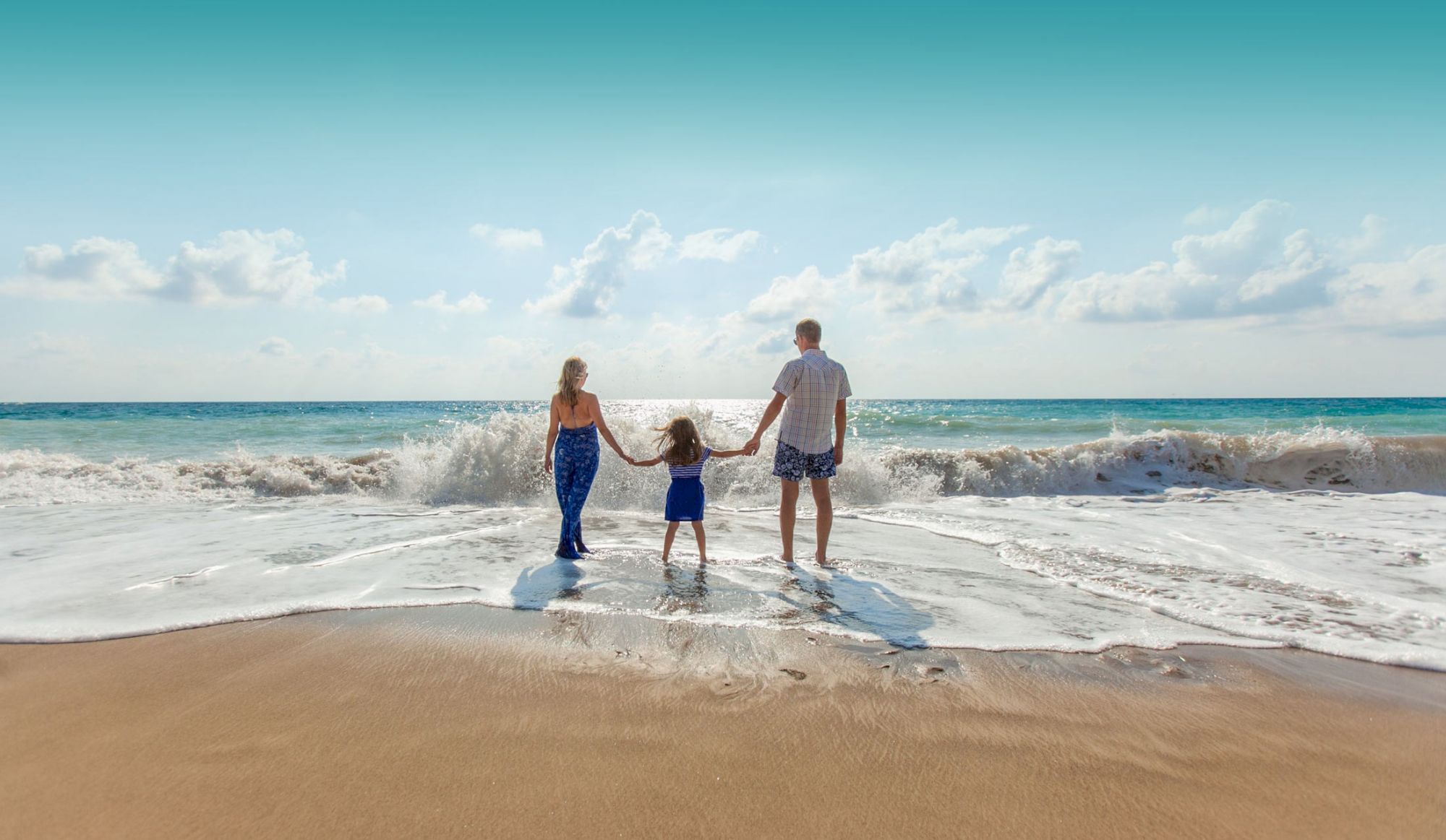 A family of three holds hands while standing on a beach, facing the ocean waves under a clear sky, enjoying a sunny day.