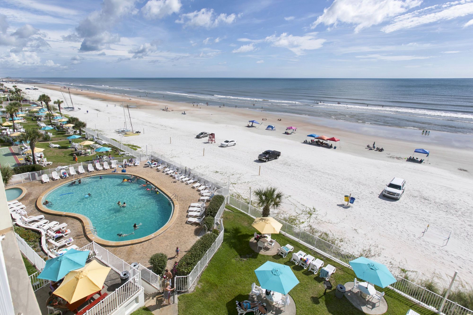 A beachfront resort with a pool, lounge chairs, and tables with umbrellas; cars and people are on the beach under a partly cloudy sky.